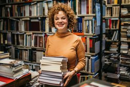 Eine Frau steht in einer Bücherei und hat einen Stapel Bücher in der Hand | © anandaBGD - Getty Images/iStockphoto