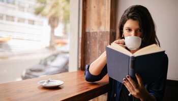 Eine Frau sitzt mit einem Kaffee am Tisch und liest dabei ein Buch | © wundervisuals - Getty Images/iStockphoto